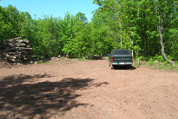 A closed-out log landing in summertime. The ground is compacted and devoid of vegetation. A pile of firewood logs remains. A green Chevy truck with a bed topper is parked on the landing.