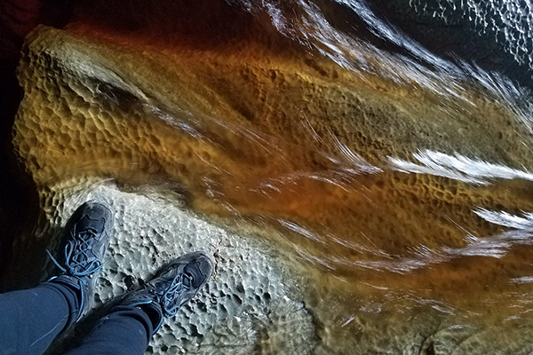 Coffee-colored water flows over bedrock with a golf ball like texture. The photographer’s boots nearly touch the water.