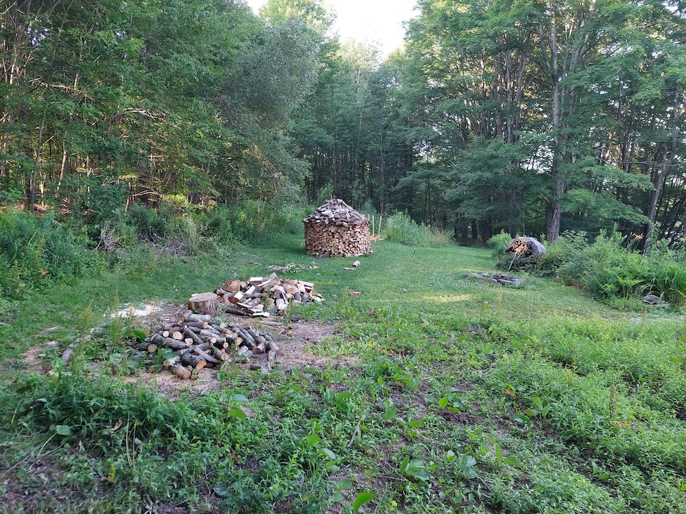 Firewood processing area. Firewood processing area showing a pile of small tree branch rounds yet to be split, a chopping block, and some split firewood. A rotund holz hausen, or wood house, is featured in the background and represents all the wood from the larger maple logs.