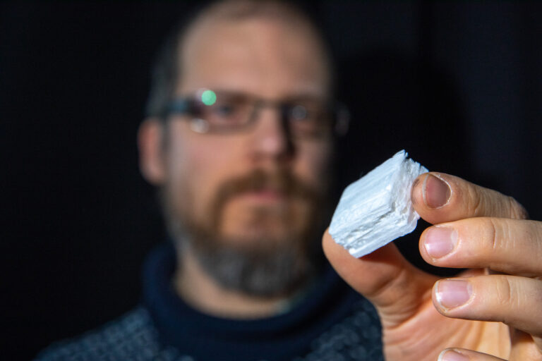 A photo of a man holding a small cube of wood foam in his thumb and index finger. 
