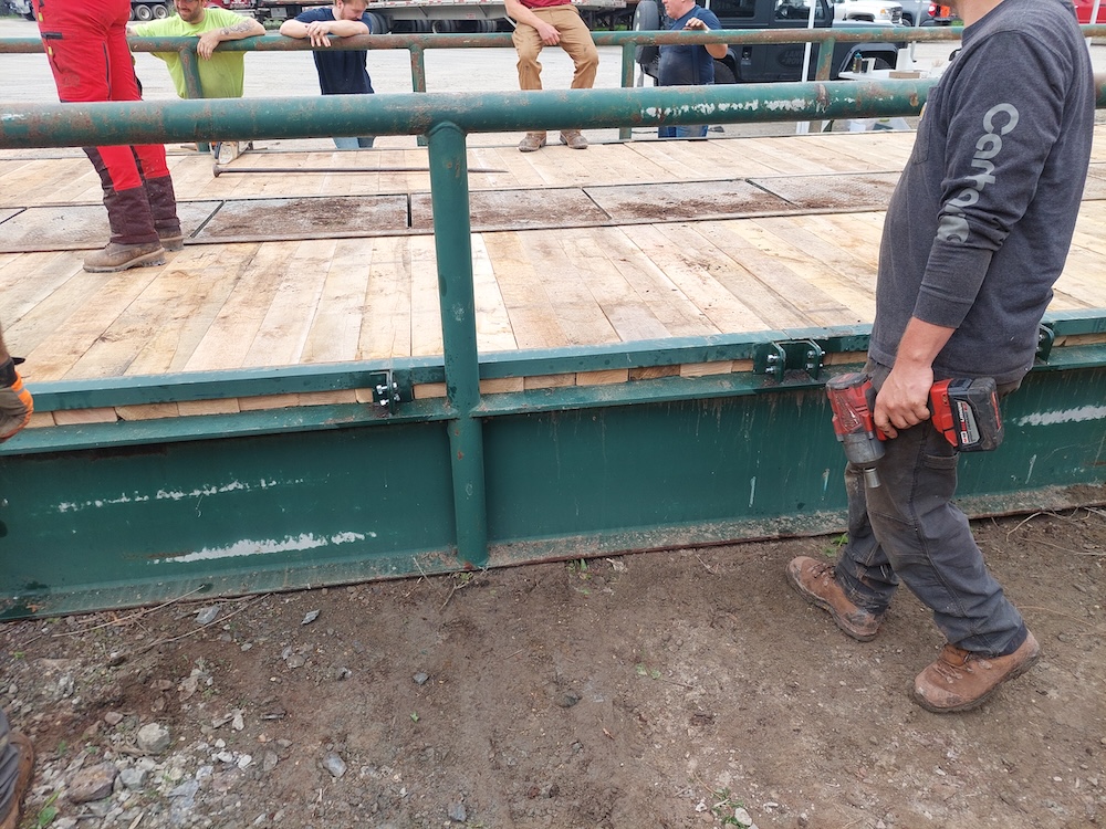 A man walks holding an impact wrench. New nuts and bolts are loosely in place. In the background, men are hanging out on and around the bridge after a job well done.