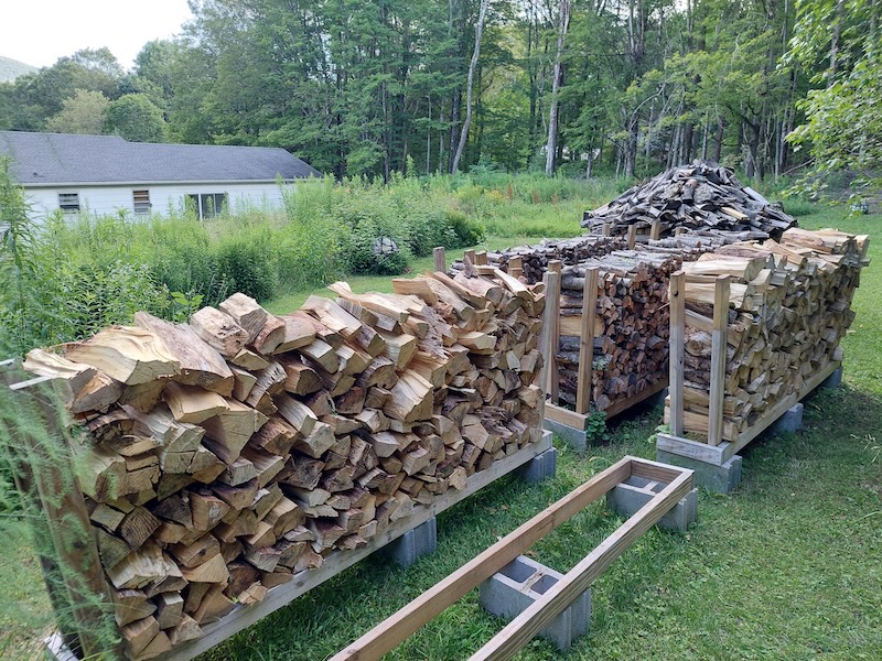 Multiple stacks of firewood in the backyard of a Catskill Mountain home during the middle of summer. The grass is neatly mowed around the firewood area with goldenrod growing wild in other areas and obscuring a white house below.