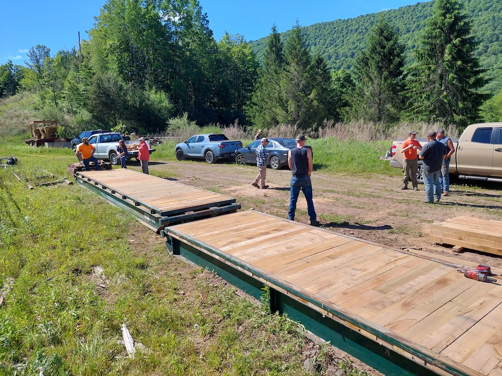 New decking, 30-foot bridge. Two 30-foot, steel-framed bridge panels lay in a construction yard with new, light-colored white oak decking. The trees have fully leafed out. It is early June. A group of forestry professionals shoot the breeze after a job well done.