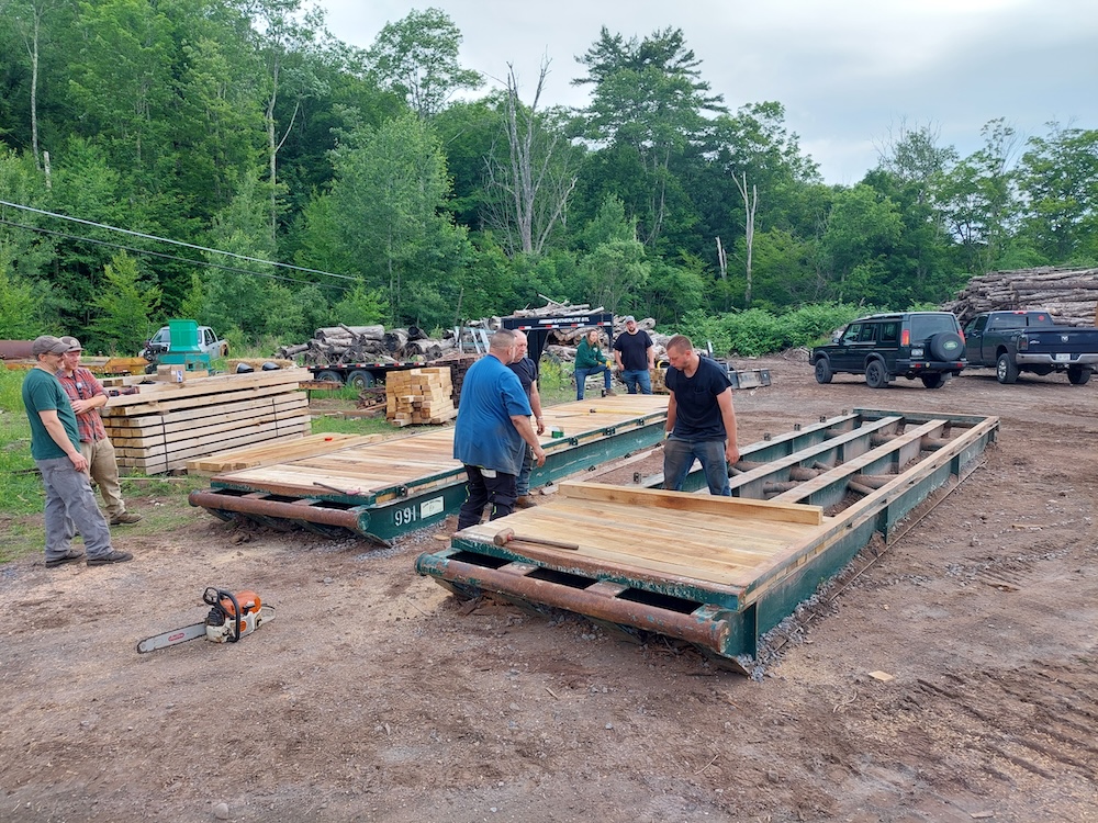New decking, second 30-foot bridge. Workers install cut-to-length lumber in a metal bridge frame at a construction yard. One 30-foot bridge panel is already complete with new decking. The panel in the foreground is one-fourth finished. 3 people are working, 4 are looking on in the background.