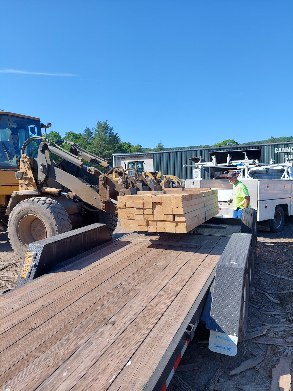 Loading the bridge decking timbers. An operator of a front-end loader with forks lowers a packet of about 50 bridge decking timbers onto a flatbed trailer while a man in high vis green gives directions. The sign on the building in the back says Cannonsville Lumber.