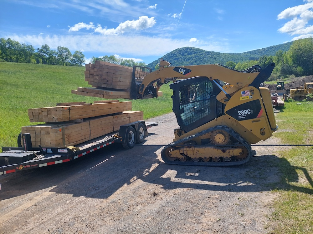 Unloading the bridge decking timbers. An operator in a Caterpillar 289C2 skid steer unloads a packet of bridge decking timbers from a flatbed trailer. There are 3 packets left on the trailer.