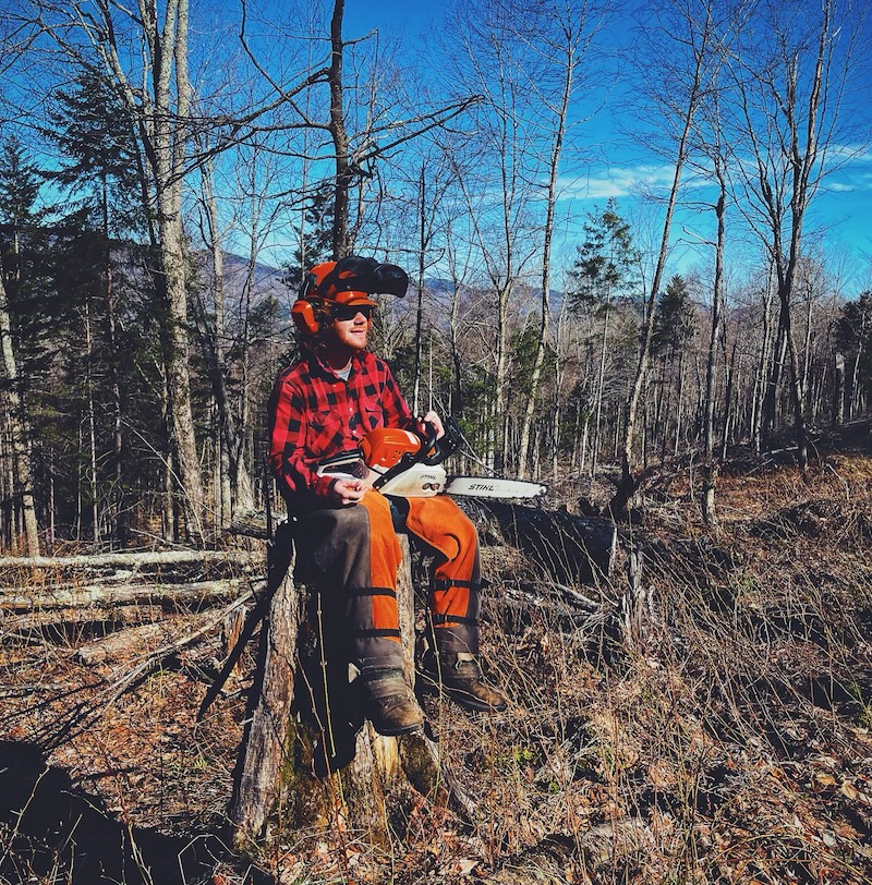 Taking a break from felling Image showing a man sitting on a tree stump holding a chainsaw in an area with felled trees wearing a hard hat, glasses, ear protection, chainsaw chaps.